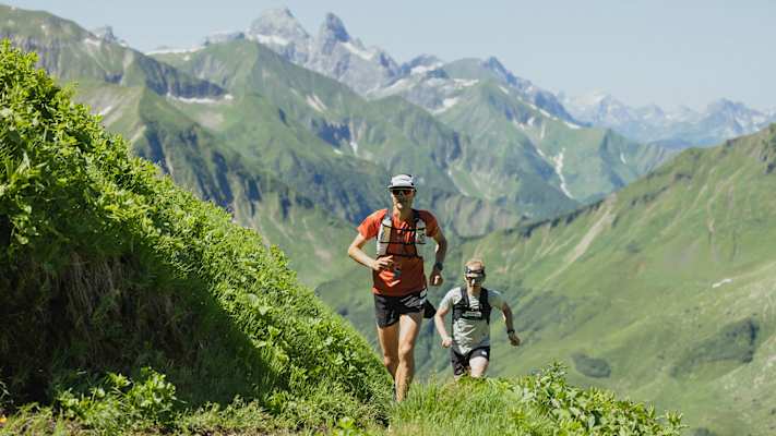 Zwei Trailrunner auf einem Bergpfad in Richtung Himmeleck, umgeben von grünen Hängen und den Gipfeln der Allgäuer Alpen.