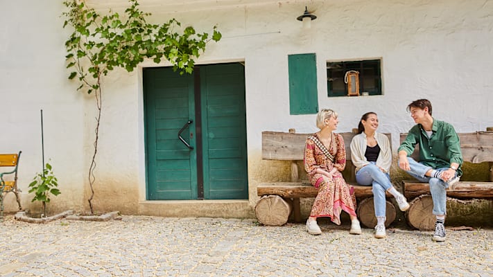 Zwei Frauen und ein Mann sitzen auf einer Holzbank vor einem Haus mit einer grünen Tür