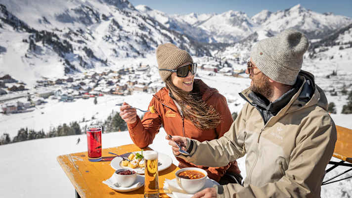 Eine Frau und ein Mann essen auf einer Hütte. Hinter ihnen ein ist ein Bergpanorama.
