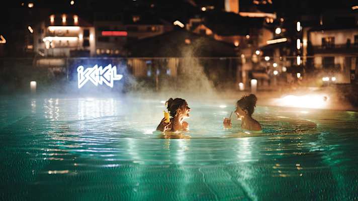 Zwei Frauen stehen im Thermalwasser in der Silvretta Therme und genießen jeweils ein Getränk.