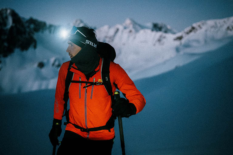 Ein Mann geht im Schnee und Skiausrüstung in der Nacht mit einer Stirnlampe skifahren.