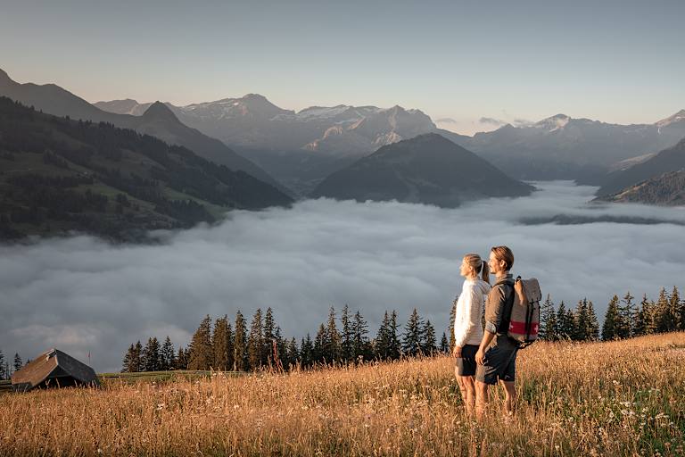 Zwei Wanderer genießen den Blick über ein Nebelmeer im herbstlichen Gstaad mit Bergpanorama im Hintergrund.