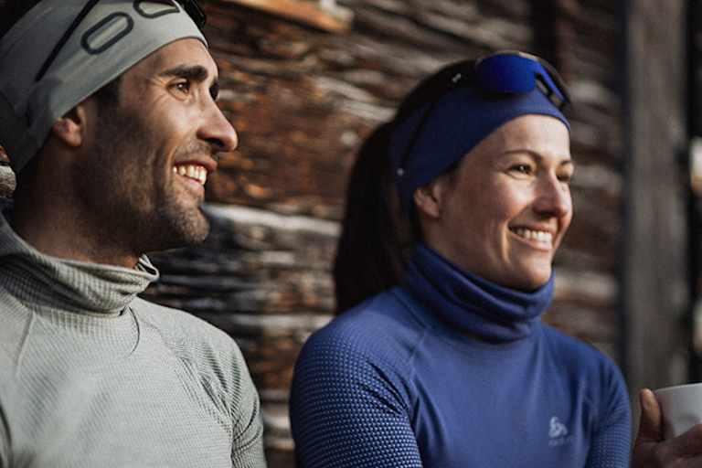 Zwei Personen in funktionaler Winterbekleidung von Odlo, genießen eine Pause im Freien vor einer rustikalen Holzhütte.