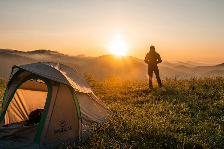 Ein Mann steht vor seinem Zelt und schaut zu wie die Sonne hinter den Bergen untergeht.