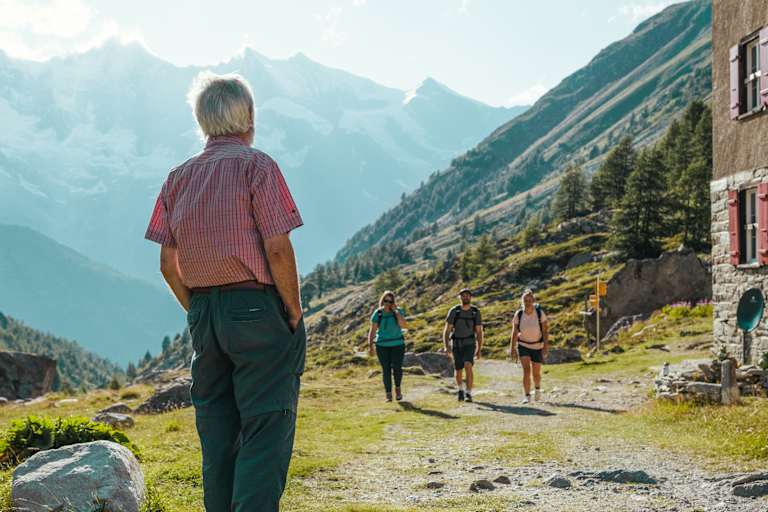 Ein Mann steht vor einer Hütte und wartet auf zwei Wanderinnen und einen Wanderer.