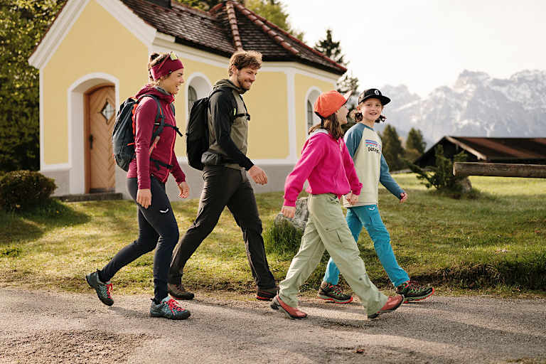 Eine Frau wandert mit einem Mann und zwei Kindern lächelnd durch eine bergige Landschaft im Frühling.