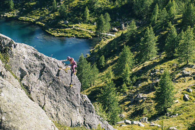 Eine Frau klettert auf einer Felswand entlang, im Hintergrund ist eine sommerliche, grüne Landschaft mit einem See zu sehen.