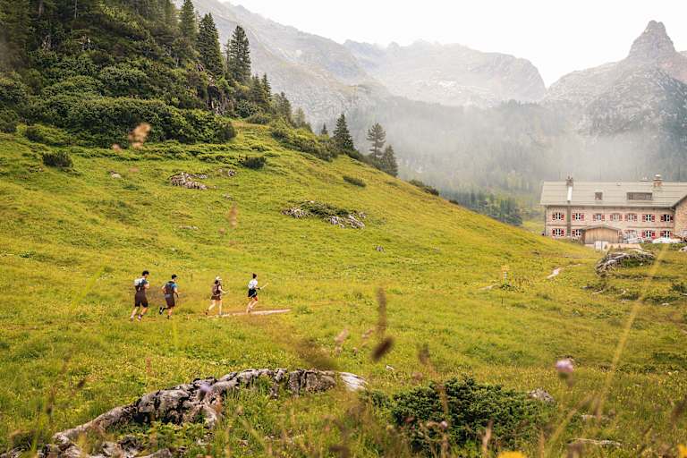 Vier Trailrunner laufen entlang einer Wiese auf einem Berg in Richtung einer Hütte mit roten Fensterläden.