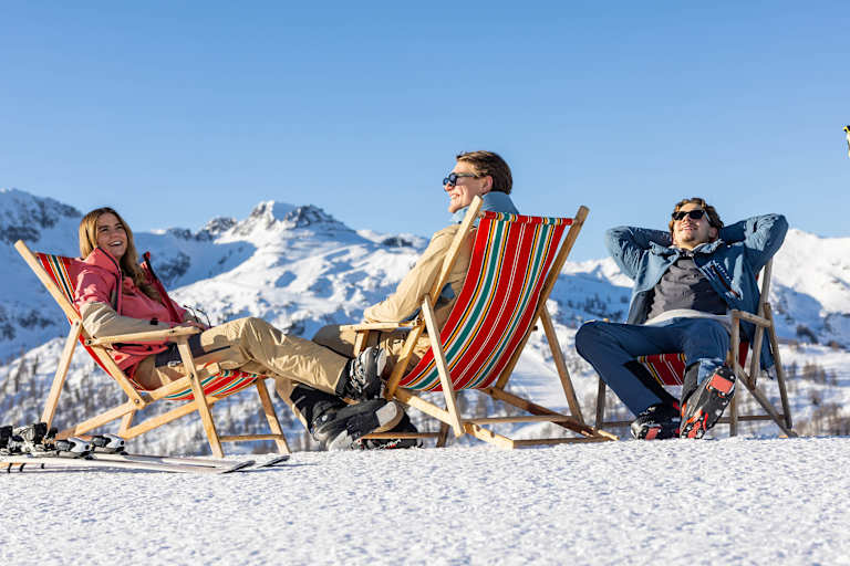 Eine Frau und zwei Männer sitzen in roten Liegenstühlen auf einem verschneiten Berg und genießen die Sonnestrahlen.