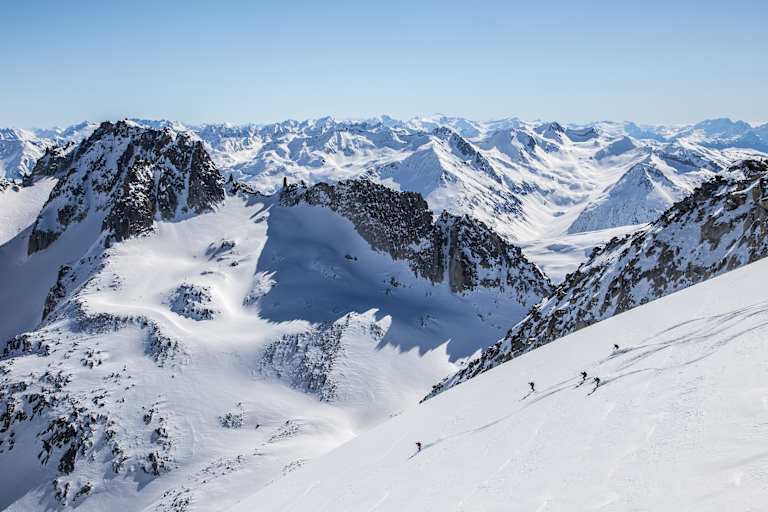 Markante Felsen die vom Schnee bedeckt sind. Skitourengeher fahren einen unverspurten Hang hinunter.