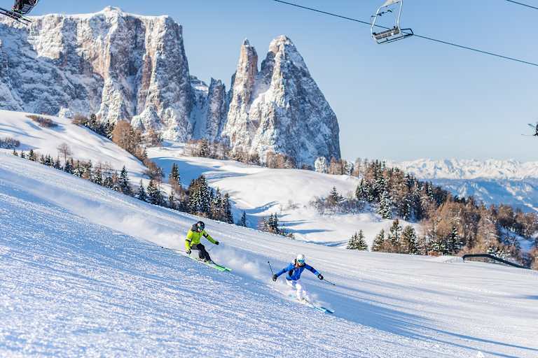Zwei Skifahrer fahren eine frisch präparierte Piste in der Dolomitenregion Seiser Alm hinunter. Im Hintergrund ist das verschneite Bergpanorama zu sehen.