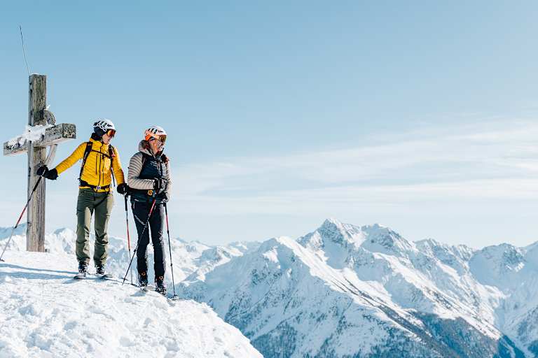 Eine Frau und ein Mann stehen neben dem Gipfelkreuz auf Tourenskiern und blicken über die verschneite Berglandschaft.