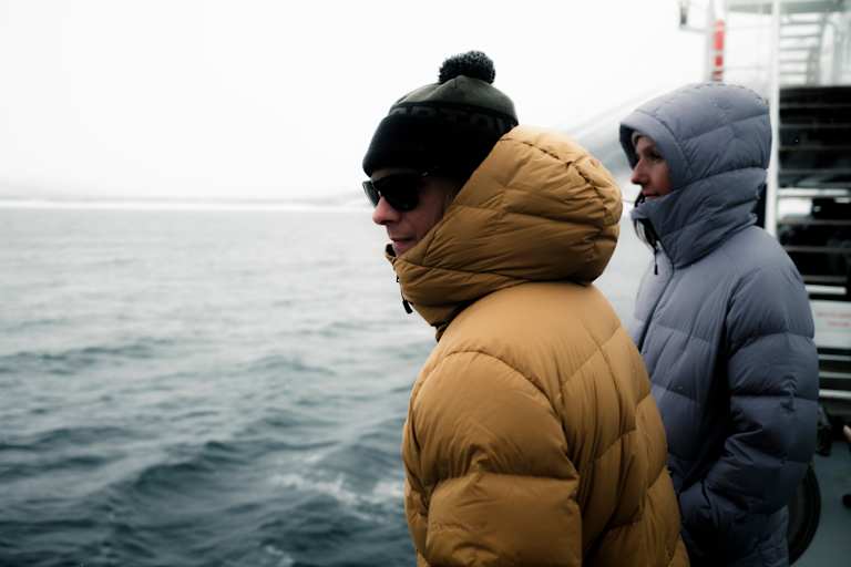 Zwei Personen stehen in Daunenjacken auf einem Schiff - hinter ihnen das Meer