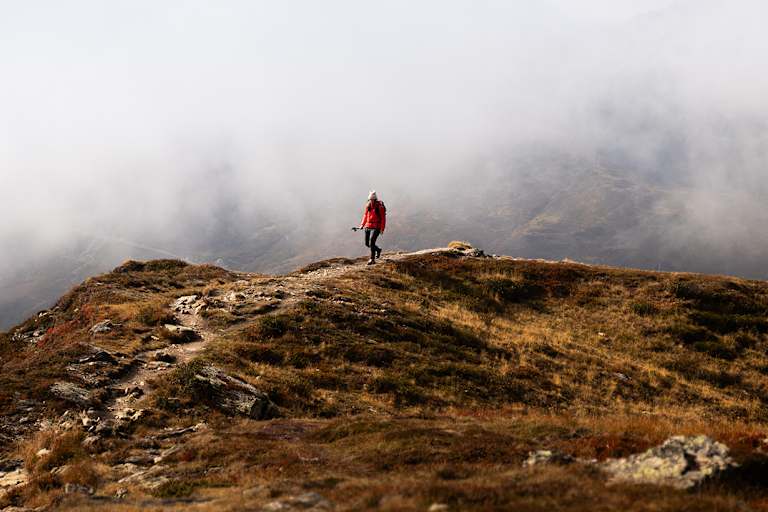 Person wandert in roter Outdoor-Jacke auf einem Bergpfad durch eine neblige Gebirgslandschaft.