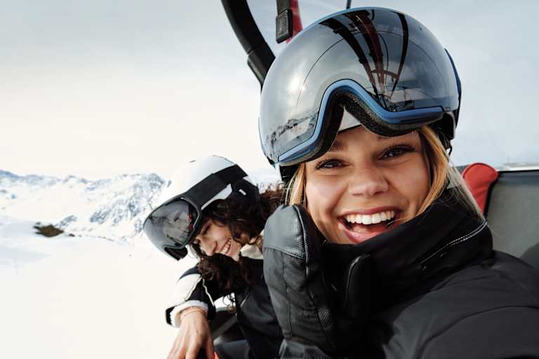 Eine Frau mit Skibrille und Helm auf dem Kopf sitz in einem Sessellift und lacht uns entgegen. Neben ihr sitzt eine weitere lächelnde Frau.
