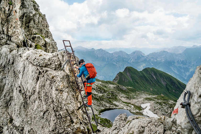 Ein Kletterer nimmt die Stahlleiter, im Hintergrund ist ein Bergsee zu sehen