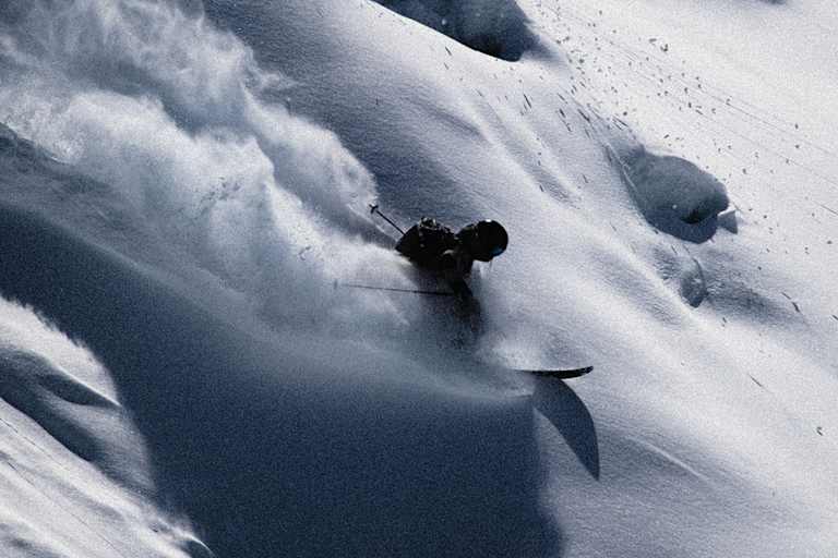 Skifahrer gleitet dynamisch durch unberührten Tiefschnee, wirbelt Pulverschnee auf und meistert eine steile Abfahrt in alpiner Winterlandschaft.
