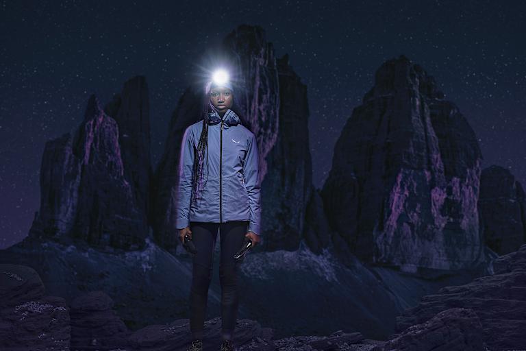 Frau mit Stirnlampe in Speedhiking Outfit von Salewa in der Nacht, im Hintergrund Berge in der Dunkelheit.