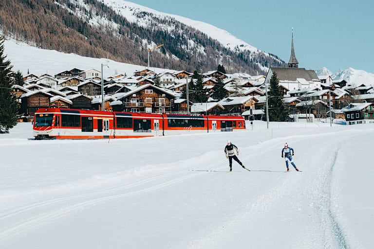 Ein Tal mit 2 Langläufern, einer Bahn und verschneite Tannen.