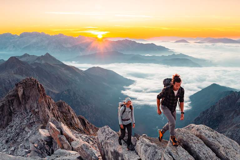Zwei Wanderer steigen bei Sonnenaufgang auf einen Dolomitengipfel im Trentino. Im Tal ein Nebelmeer.