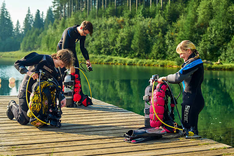 Drei Taucher in Neoprenanzügen knien auf einem Holzsteg am Grüblsee