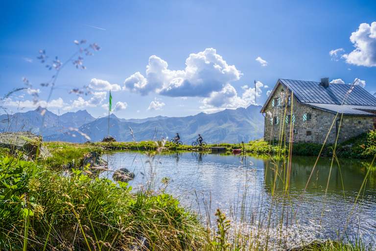 Zwei Mountainbiker radeln an einem Bergsee entlang. Daneben steht eine Hütte.
