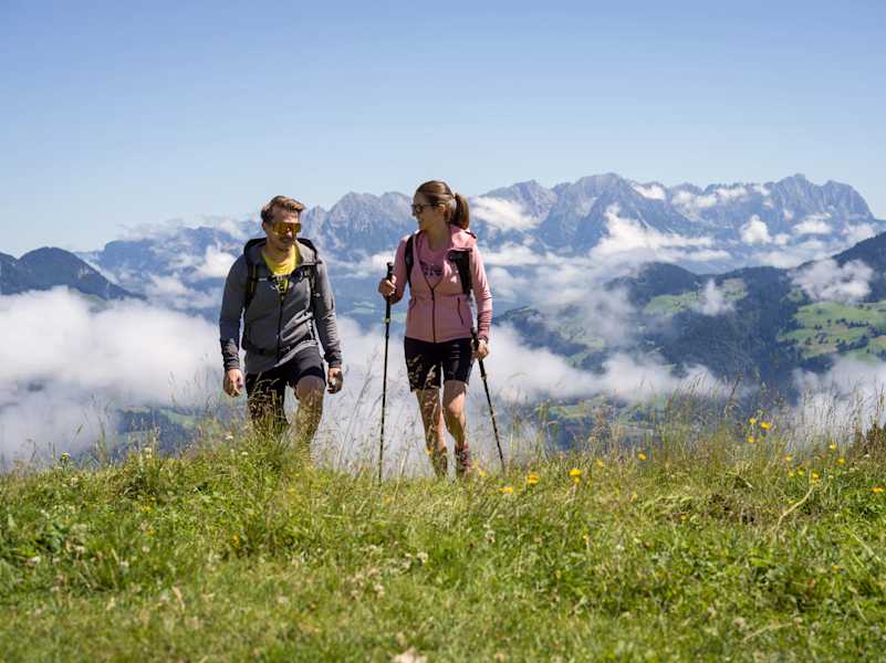 Ein Mann und eine Frau wandern über eine Wiese, hinter ihnen ein Bergpanorama.