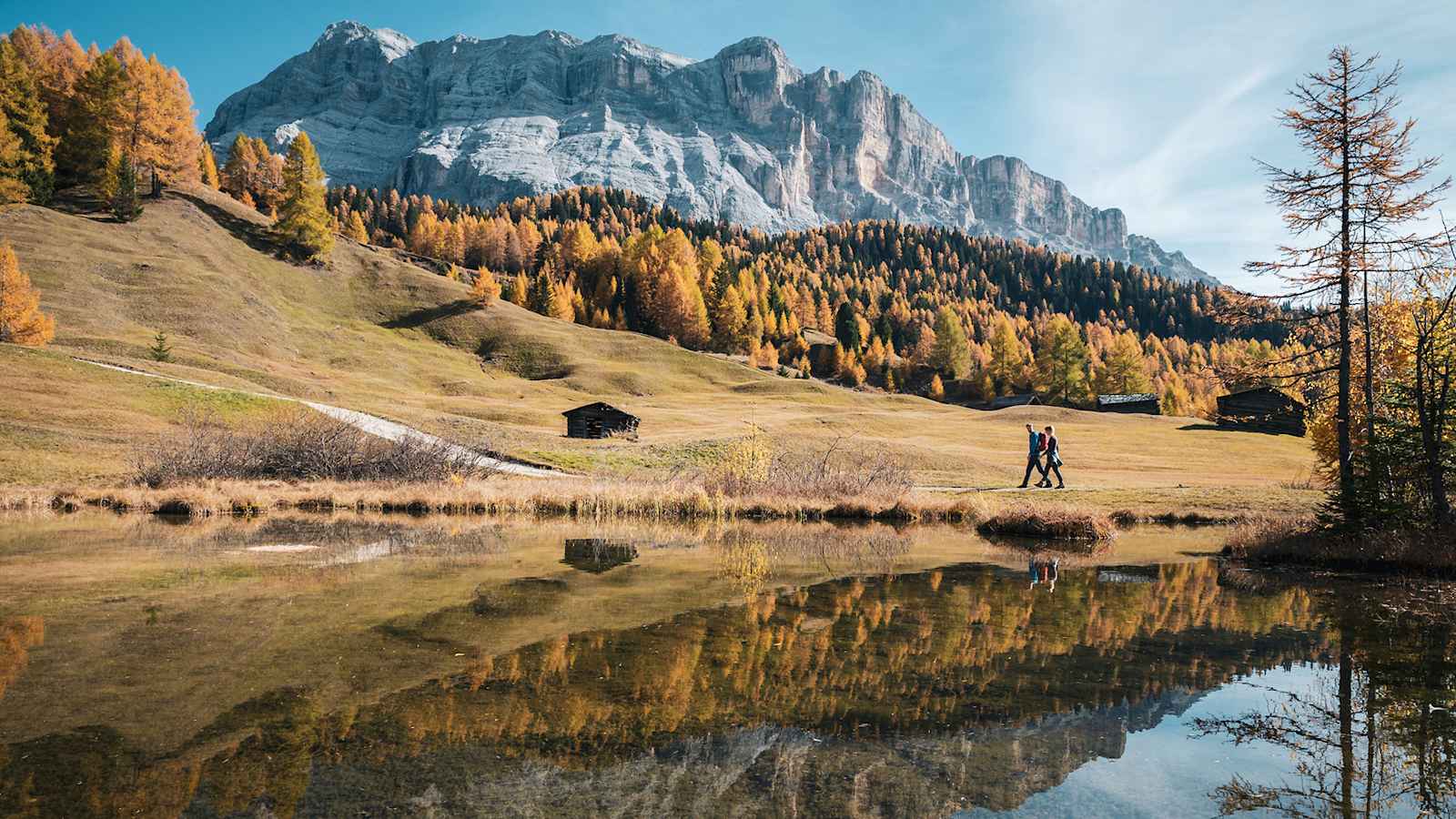 Zwei Wanderer gehen an einem See vorbei, in dem sich die Dolomiten spiegeln.