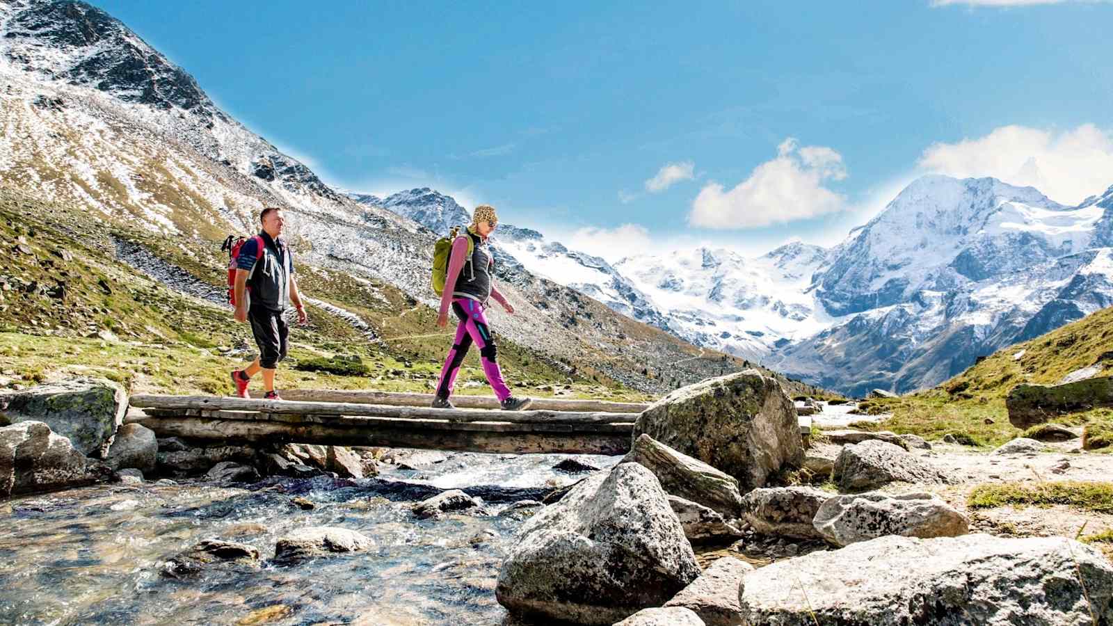 Wanderer überqueren einen Gebirgsbach, schneebedeckte Berge im Hintergrund