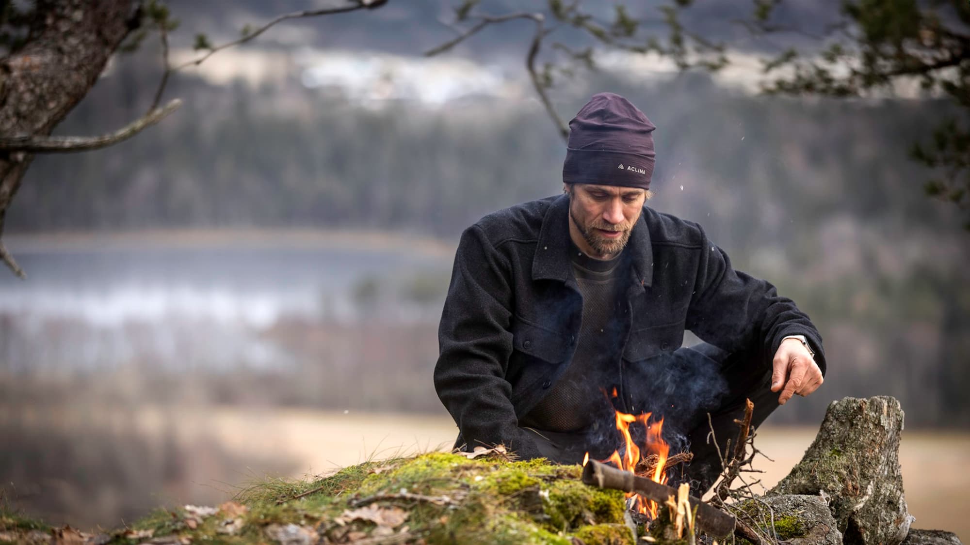 Ein Mann mit Haube und Jacke von Aclima macht ein Lagerfeuer in der Natur.