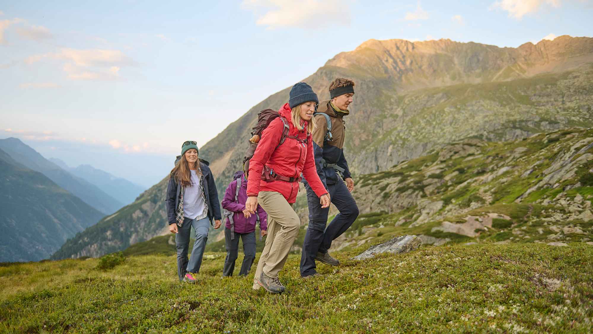 Eine Gruppe von Wanderern in funktionaler Outdoor-Bekleidung bewegt sich durch eine malerische Berglandschaft. Grüne Wiesen und majestätische Gipfel bilden eine beeindruckende Kulisse.