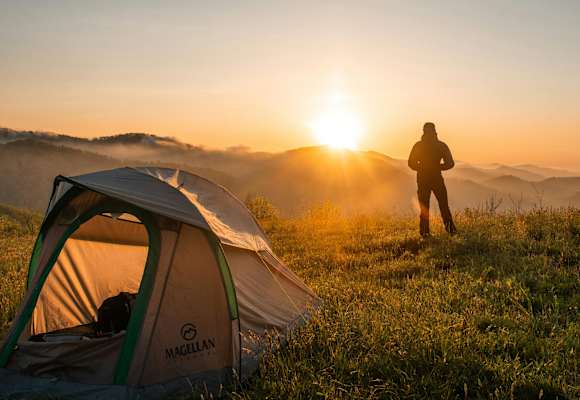 Ein Mann steht vor seinem Zelt und schaut zu wie die Sonne hinter den Bergen untergeht.