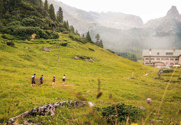 Vier Trailrunner laufen entlang einer Wiese auf einem Berg in Richtung einer Hütte mit roten Fensterläden.