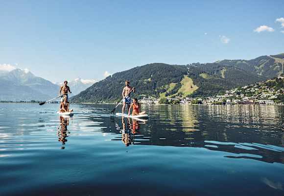 Zwei Männer tauche mit einem Puddle ihr Stand-up-paddeln an. Jeweils eine Frau, sitz am vorderen Ende. Sie gleiten über denn Zeller See. Hinter ihnen ein Bergpanorama.
