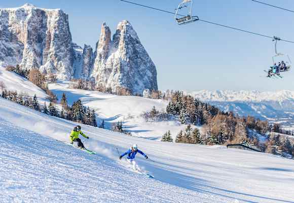 Zwei Skifahrer fahren eine frisch präparierte Piste in der Dolomitenregion Seiser Alm hinunter. Im Hintergrund ist das verschneite Bergpanorama zu sehen.