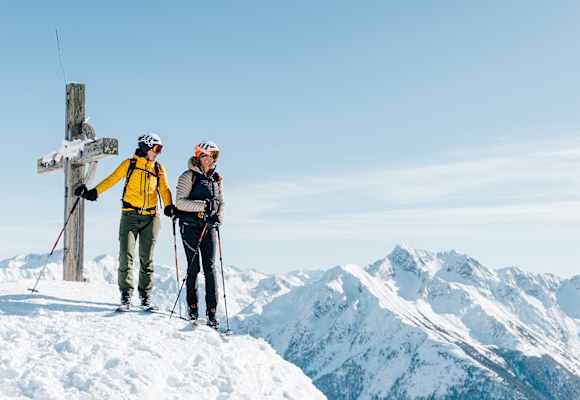 Eine Frau und ein Mann stehen neben dem Gipfelkreuz auf Tourenskiern und blicken über die verschneite Berglandschaft.