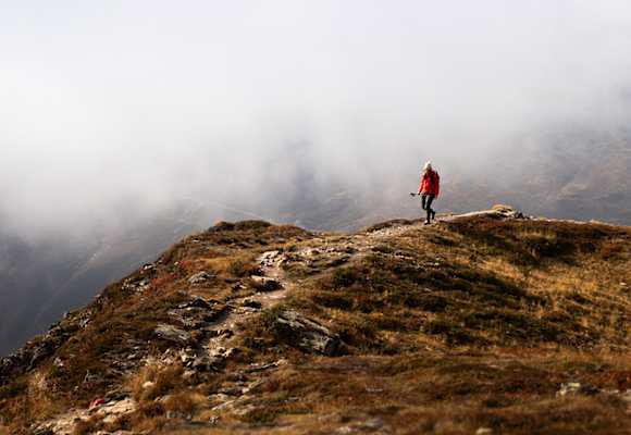 Person wandert in roter Outdoor-Jacke auf einem Bergpfad durch eine neblige Gebirgslandschaft.