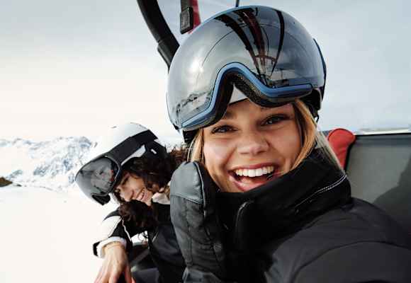 Eine Frau mit Skibrille und Helm auf dem Kopf sitz in einem Sessellift und lacht uns entgegen. Neben ihr sitzt eine weitere lächelnde Frau.