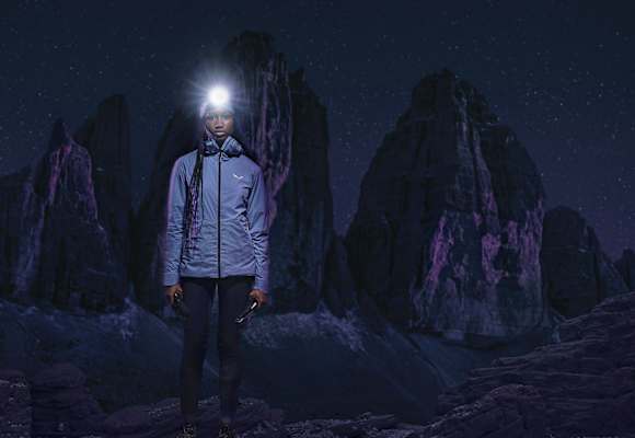 Frau mit Stirnlampe in Speedhiking Outfit von Salewa in der Nacht, im Hintergrund Berge in der Dunkelheit.