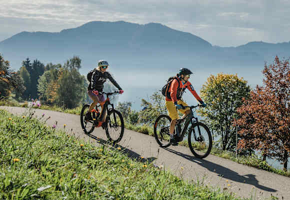 Zwei Radfahrer auf einer Straße in Oberösterreich, im Hintergrund Berge und buntgefärbte Bäume.